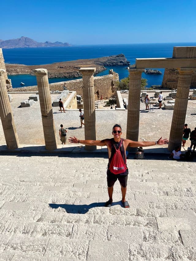 Un uomo in un viaggio di gruppo WeRoad posa a braccia aperte sui gradini in pietra di antiche rovine che si affacciano sul mare.