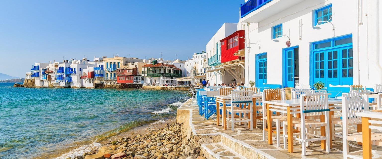 Una terrazza di ristorante sul lungomare con mobili blu e bianchi si trova accanto a un mare turchese e a edifici costieri imbiancati a calce.