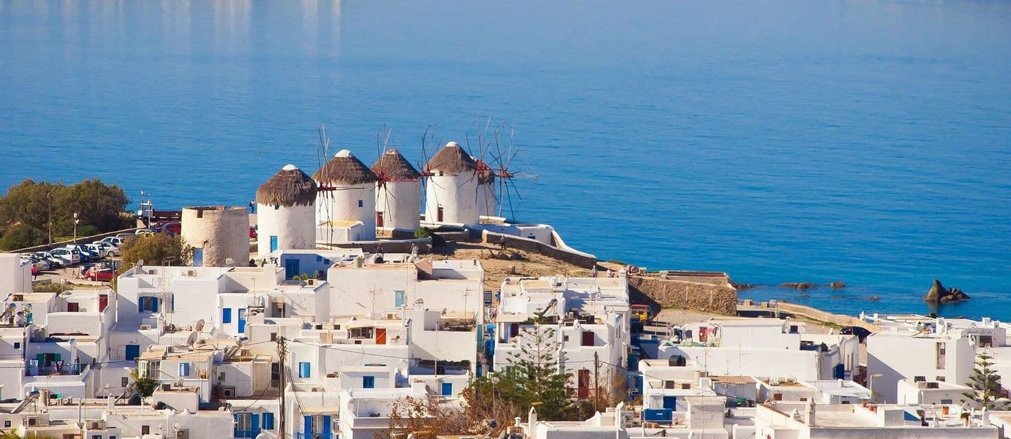 Cittadina costiera imbiancata a calce e tradizionali mulini a vento con tetti di paglia, affacciati su un mare blu e calmo.