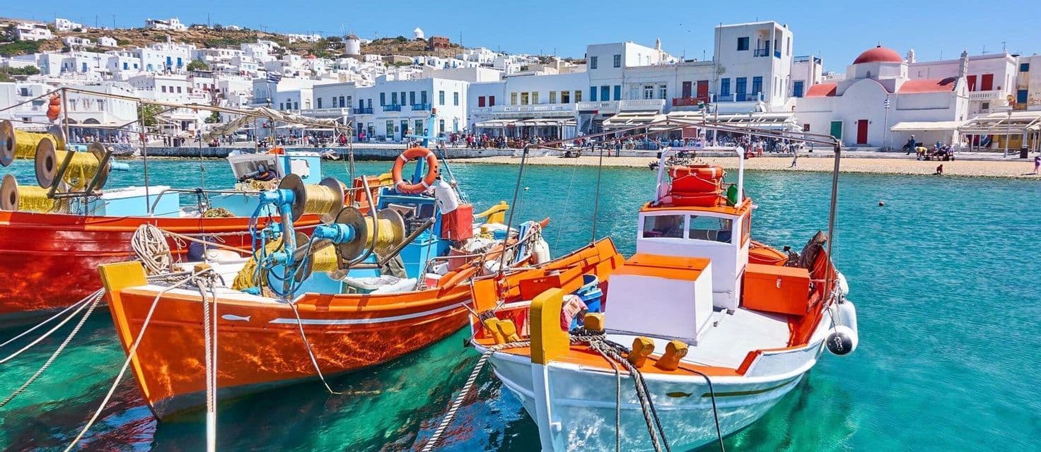 Pescherecci colorati ormeggiati in un porto con acque limpide e turchesi, davanti a una città costiera con case bianche.