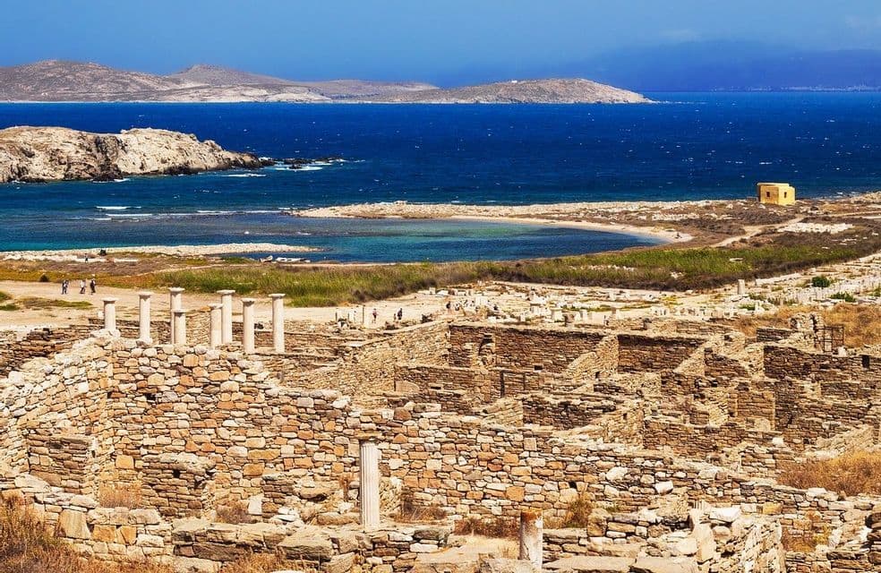 Antiche rovine di pietra con colonne si ergono su una collina che domina una baia con acque blu profonde e isole lontane.