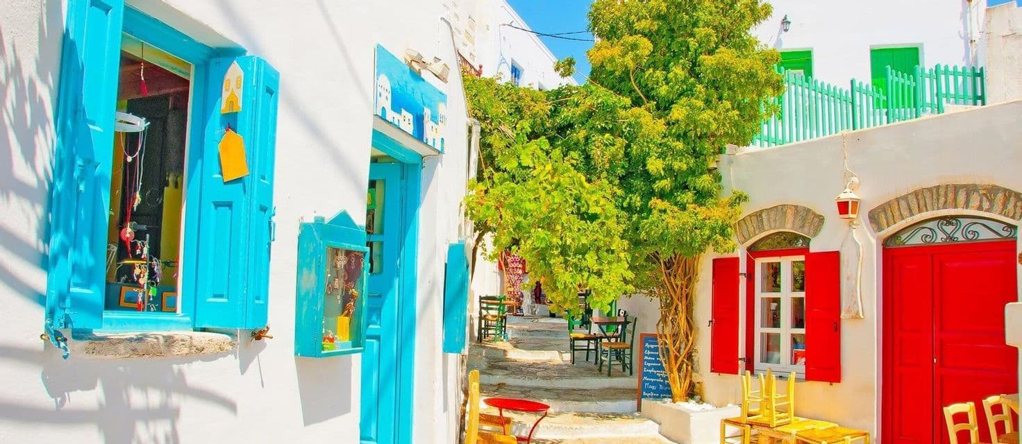 Un vicolo soleggiato con edifici imbiancati, caratterizzato da porte e persiane blu e rosse vivaci e un grande albero verde al centro.