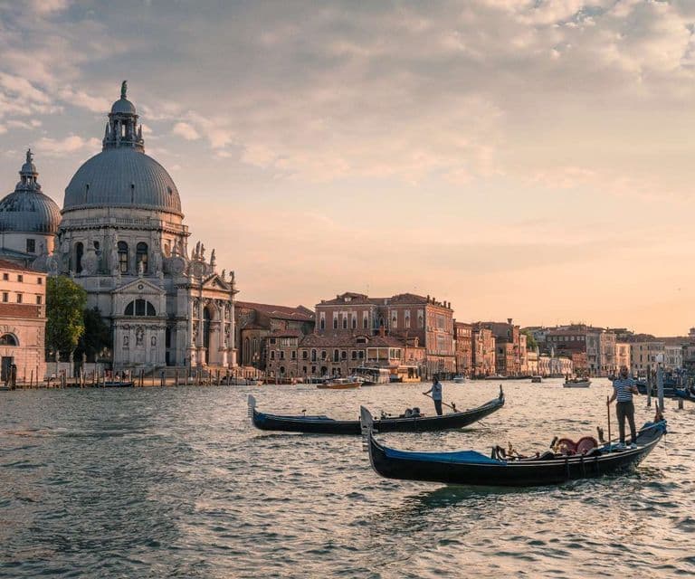 Gondole galleggiano su un canale di fronte a una grande basilica a cupola e a edifici storici al tramonto.