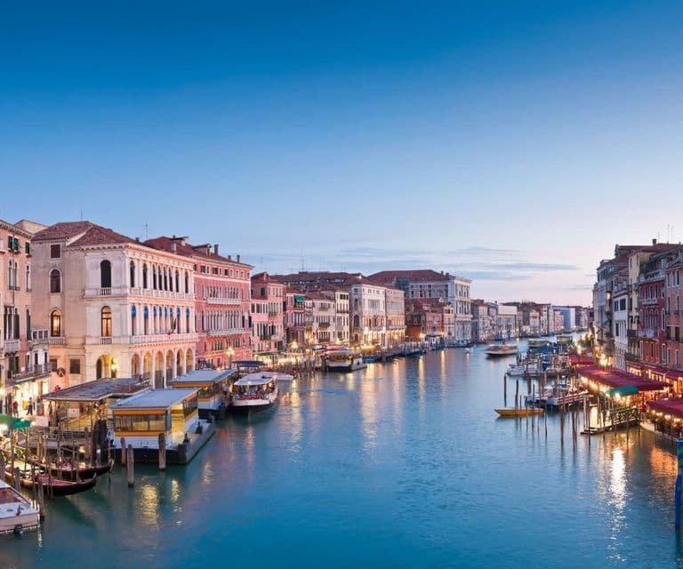 Des bâtiments historiques illuminés bordent un large canal rempli de bateaux et de vaporettos sous un ciel bleu clair au crépuscule.
