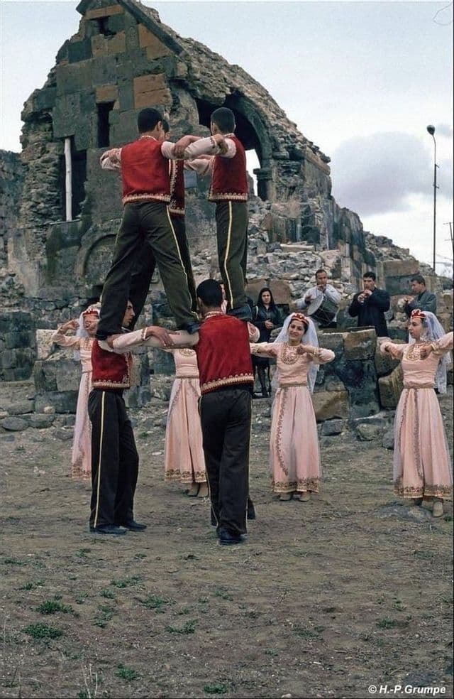 Persone in abiti tradizionali eseguono una danza popolare, con uomini che formano una piramide umana davanti a rovine di pietra.