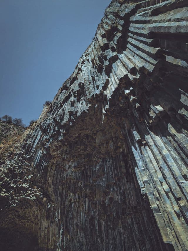 Una vista dal basso di un'enorme parete rocciosa composta da colonne di basalto esagonali, sotto un cielo azzurro limpido.