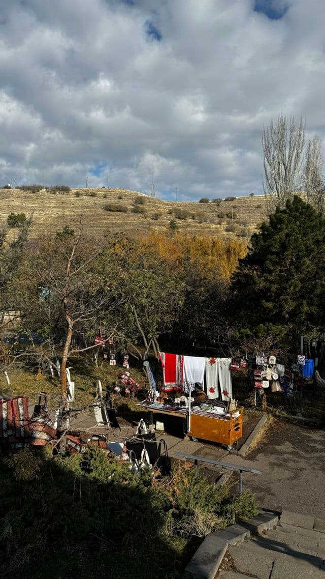 Una bancarella di mercato all'aperto con tessuti e artigianato, esposta sotto una collina erbosa e un cielo nuvoloso.