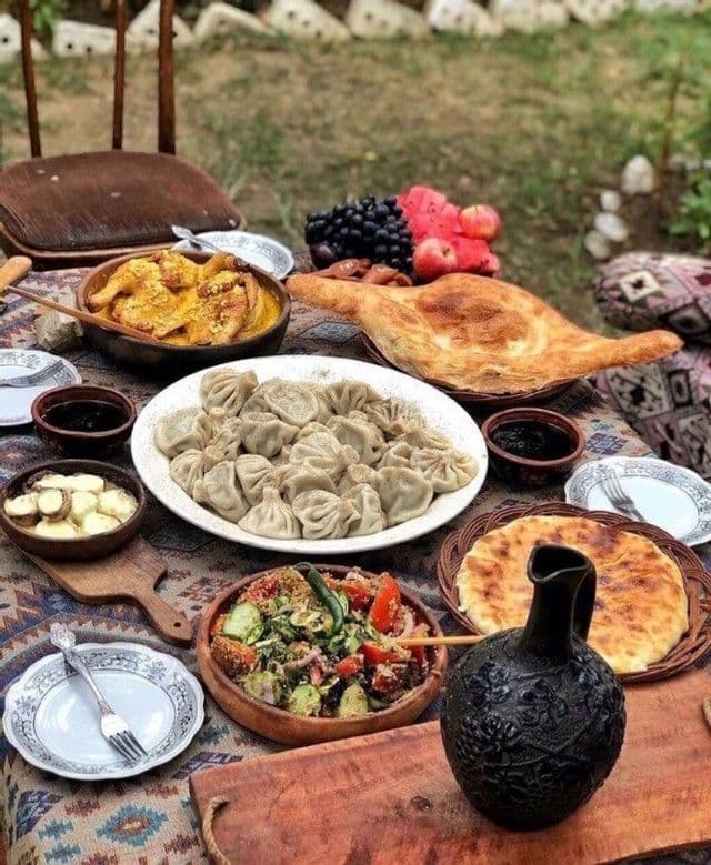 Una tavola apparecchiata all'aperto con una selezione di cibo tradizionale, inclusi gnocchi, stufato, insalata, frutta fresca e pane.