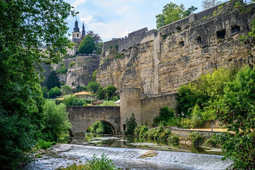 Un ponte ad arco in pietra attraversa un fiume con un piccolo stramazzo, sovrastato da storiche fortificazioni in pietra su una scogliera e con guglie di chiese in lontananza.