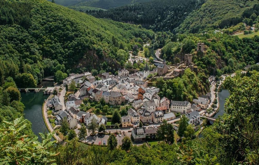 Una vista aerea di un piccolo villaggio annidato in un'ansa di fiume, con un castello in rovina su una collina, circondato da verdi montagne boscose.