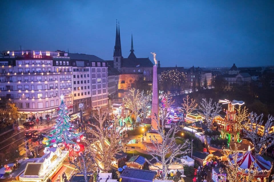 Una piazza cittadina con un mercatino di Natale splendidamente illuminato al crepuscolo, vista dall'alto, caratterizzata da un grande albero e un obelisco.