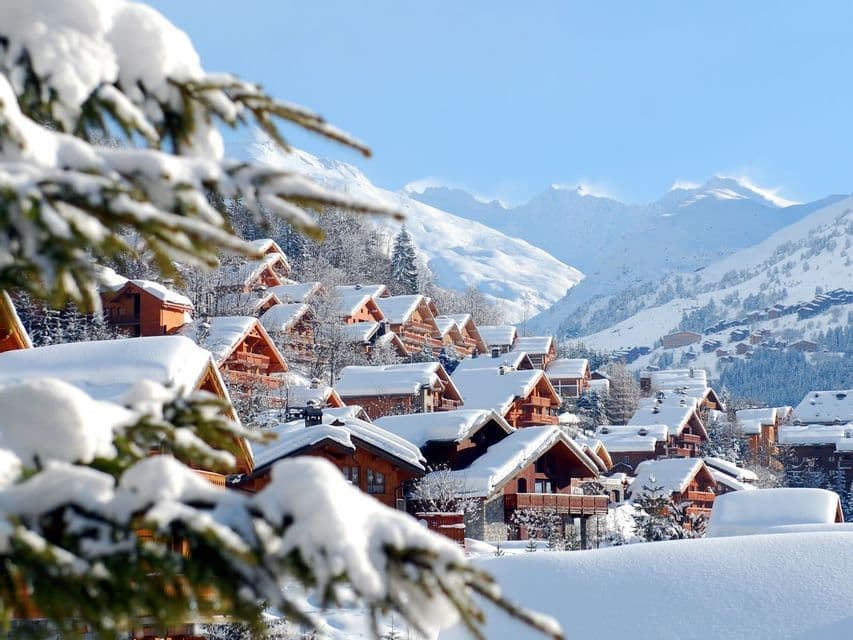 Eine Ansammlung von tief verschneiten Holzchalets an einem Berghang, mit schneebedeckten Gipfeln im Hintergrund.