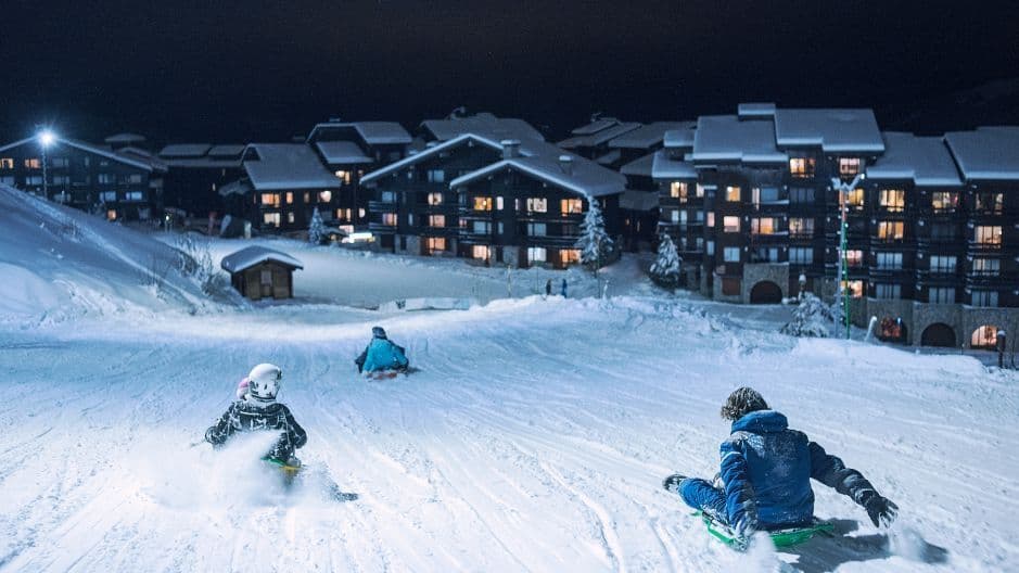 WeRoad Gruppenreise: Nachtrodeln auf verschneitem Hang mit beleuchteten Chalets im Hintergrund.