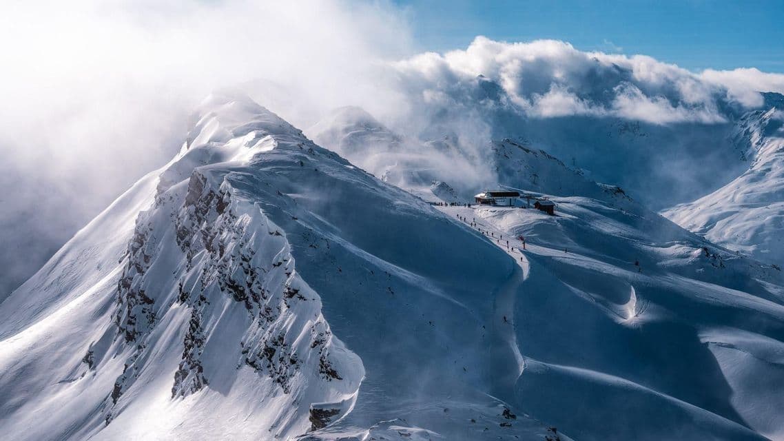 Une vaste chaîne de montagnes enneigée par une journée venteuse, avec des skieurs descendant une piste devant un chalet de montagne.