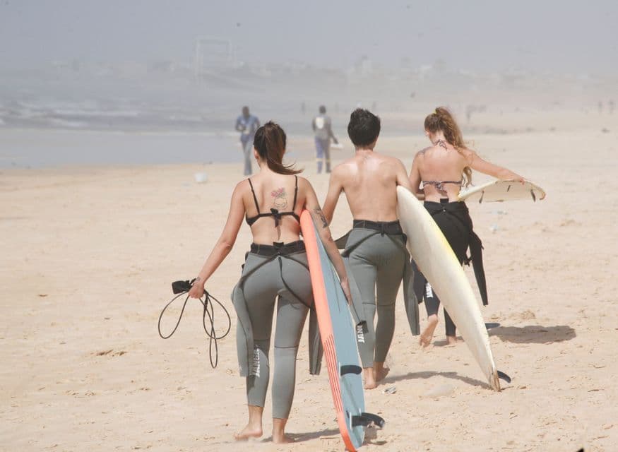 Un viaggio di gruppo WeRoad con tre surfisti in muta che camminano su una spiaggia sabbiosa con le loro tavole da surf verso l'oceano.
