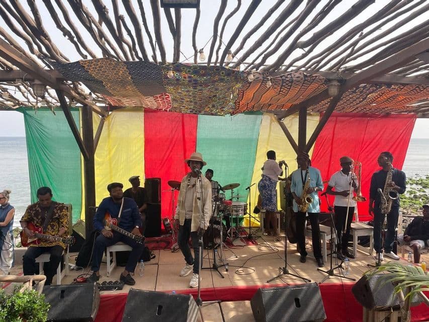 Una band con un cantante, chitarristi e strumenti a fiato si esibisce su un palco all'aperto in riva al mare sotto un pergolato in legno.