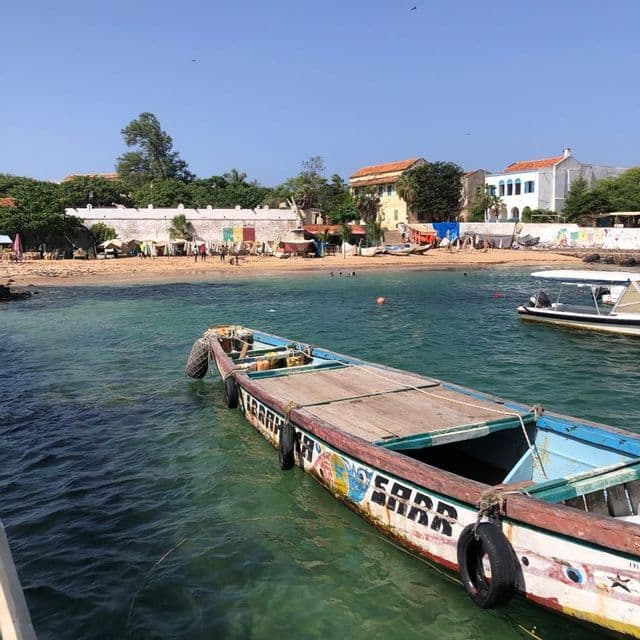 Una lunga barca di legno colorata galleggia in una baia, con una spiaggia sabbiosa, persone ed edifici visibili sulla riva.