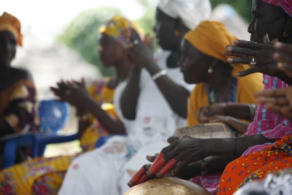 Un gruppo di donne in abiti tradizionali colorati e foulard, che suonano i tamburi e battono le mani.