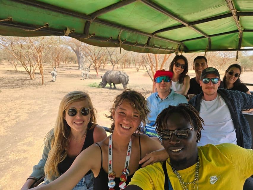 Un gruppo WeRoad sorride per una foto in un veicolo da safari, con un rinoceronte e delle zebre che pascolano sullo sfondo.