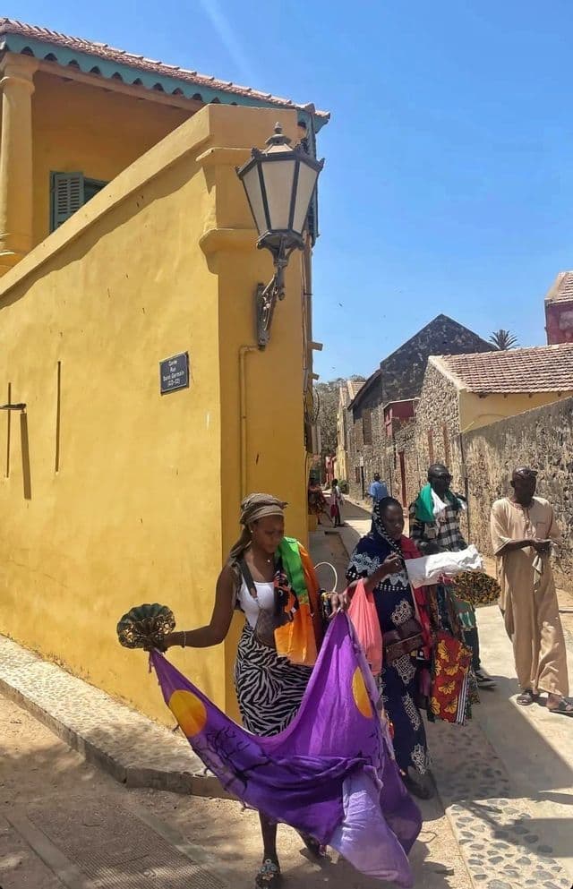 Una donna mostra un panno viola e un ventaglio su una soleggiata strada di ciottoli, mentre altre persone passeggiano accanto a un edificio giallo.