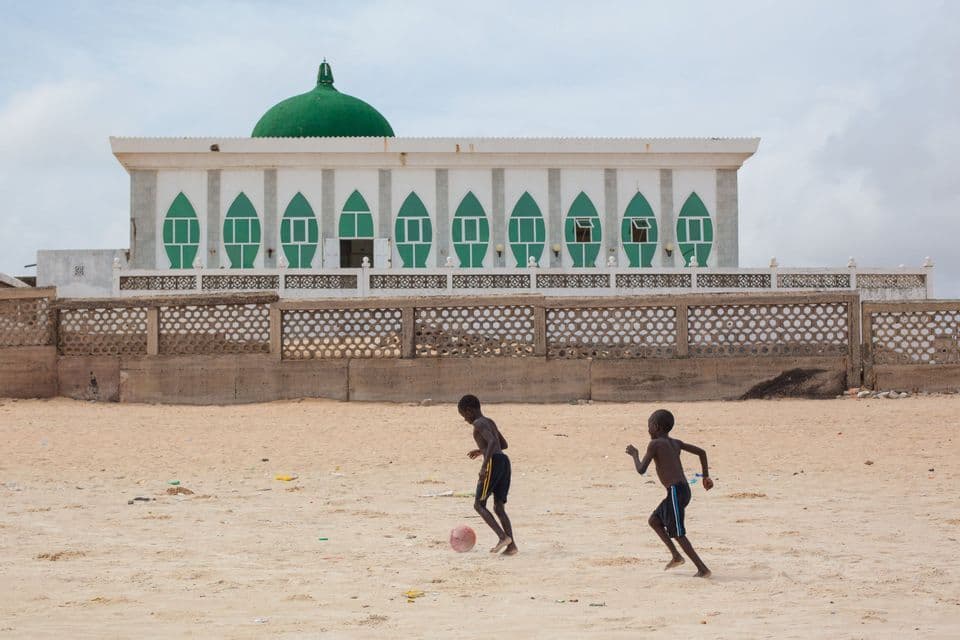 Due giovani ragazzi giocano a calcio su un campo sabbioso di fronte a una grande moschea bianca con una cupola verde.
