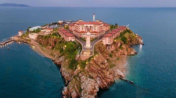 Veduta aerea di un grande e simmetrico complesso alberghiero su una penisola rocciosa circondato dal mare al tramonto.