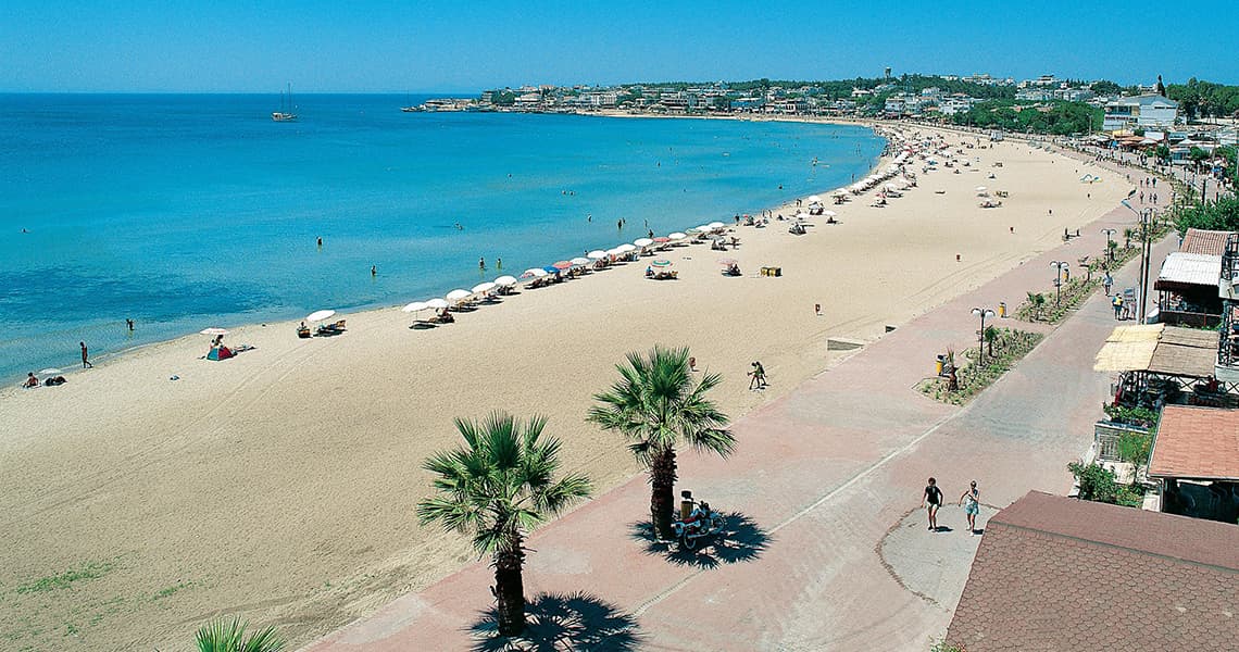 Vista dall'alto di una spiaggia assolata e affollata, con sabbia bianca, acqua turchese e un lungomare costeggiato da palme.