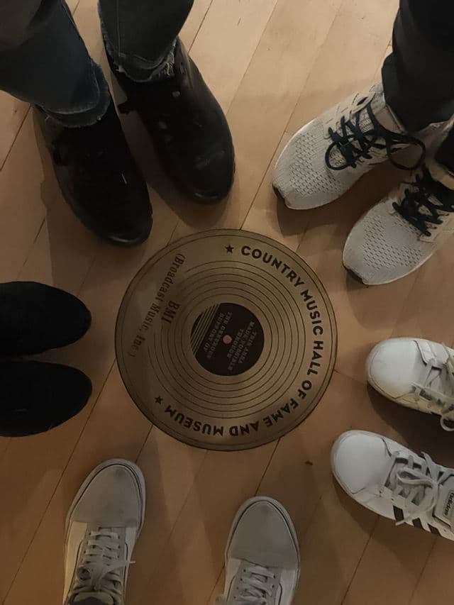 Vue d'en haut des pieds d'un groupe WeRoad réunis autour d'une plaque au sol du Country Music Hall of Fame and Museum.
