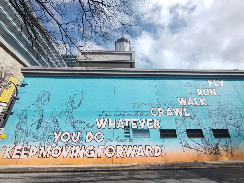 Une grande fresque murale bleue et orange sur le côté d'un immeuble, avec la citation motivante 'Continuons d'avancer'.