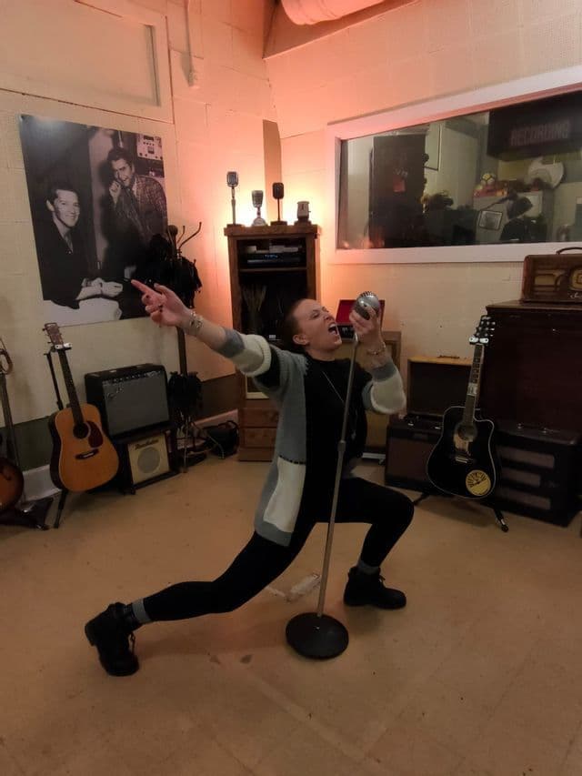 Una persona in posa canta in un microfono vintage all'interno di uno studio di registrazione musicale.