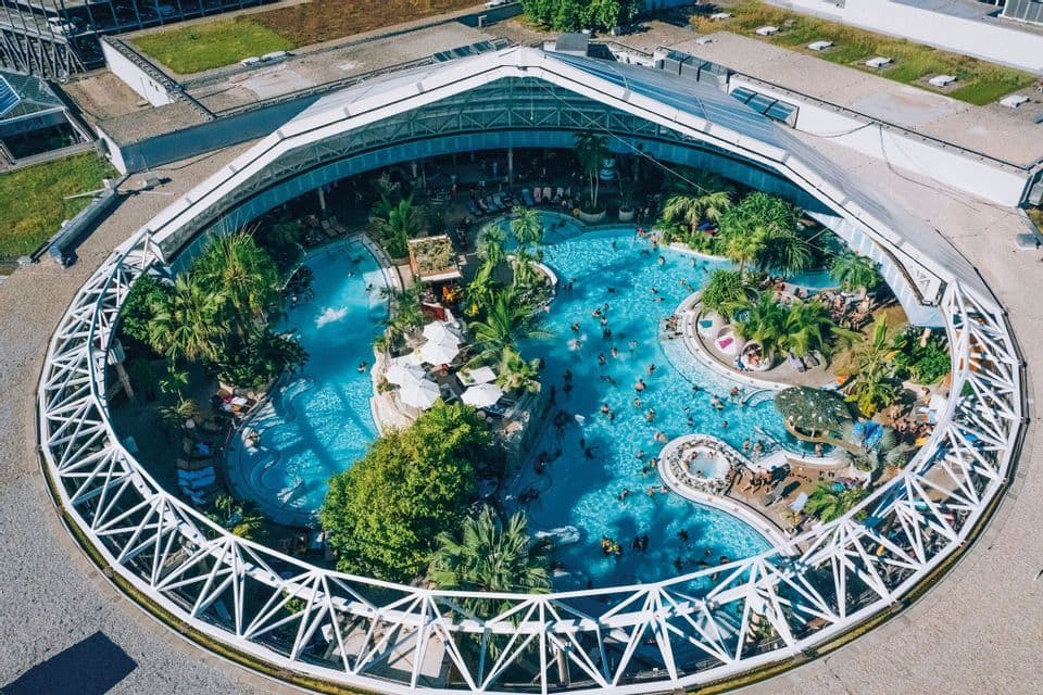 Una vista aerea di un grande complesso di piscine circolari dove le persone nuotano e si rilassano tra piante tropicali sotto un'ampia cupola.