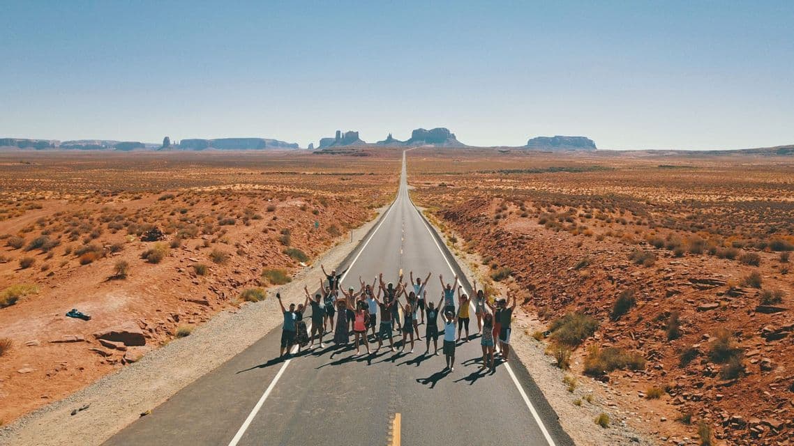 Vista aerea di un gruppo WeRoad in viaggio che posa con le braccia alzate su una lunga strada attraverso un vasto paesaggio desertico.