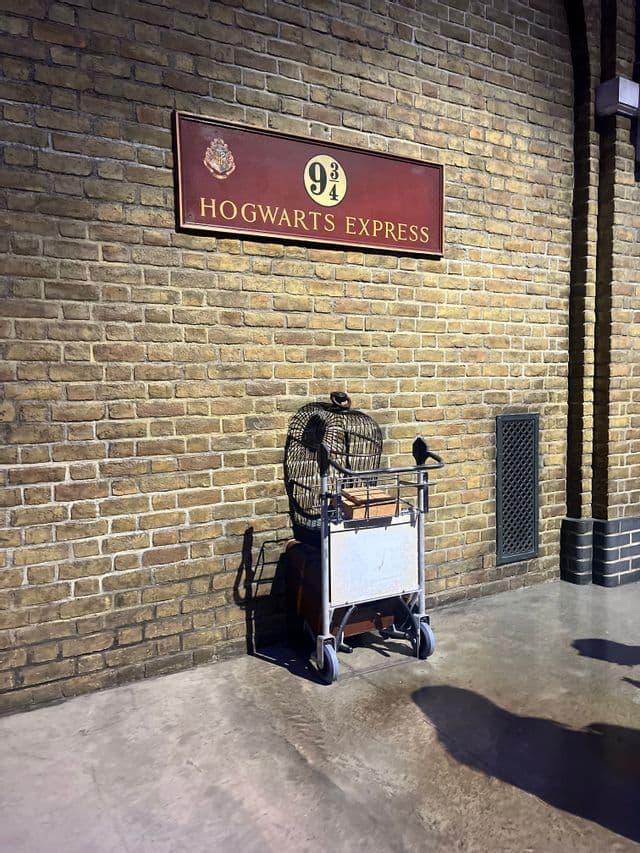 Un carrello per bagagli con una gabbia per uccelli e una valigia è posizionato a metà in un muro di mattoni sotto il cartello del Binario 9 3/4 dell'Hogwarts Express.