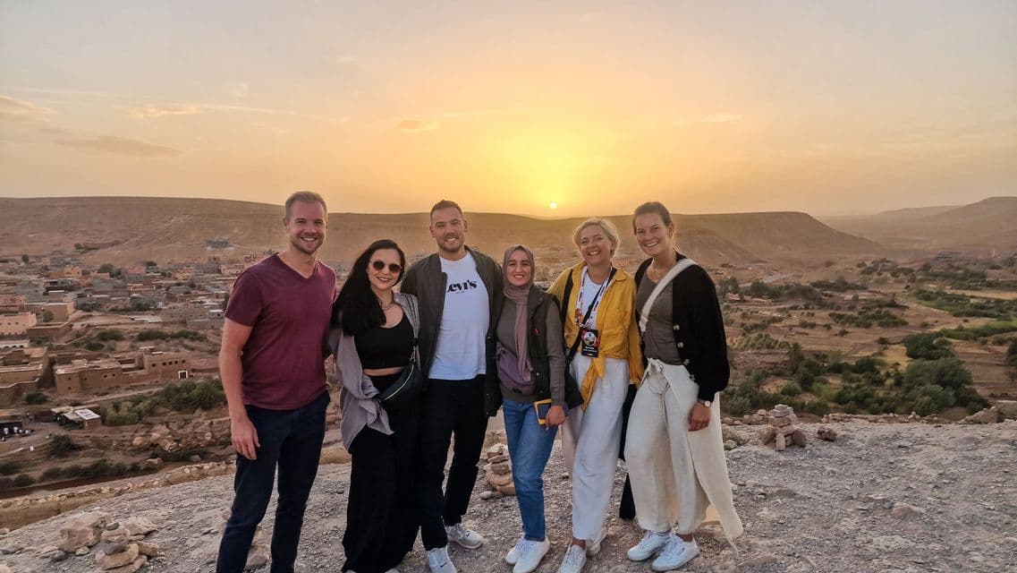 Eine sechsköpfige WeRoad-Gruppe posiert auf einem Hügel mit Blick auf ein Tal mit Lehmbauten bei Sonnenuntergang.