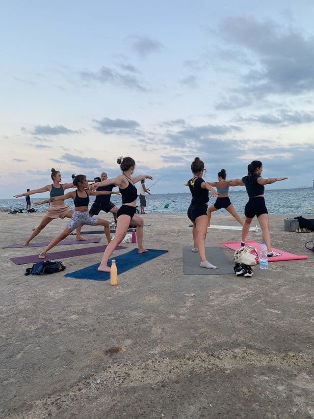 Eine WeRoad Gruppenreise, bei der Yoga in Kriegerhaltung auf Matten auf einem Betonsteg am Meer unter bewölktem Himmel praktiziert wird.