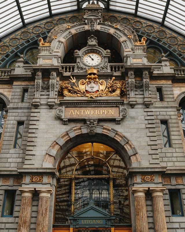 Façade en pierre ornée à l'intérieur d'un bâtiment avec une grande horloge, un blason doré et un panneau indiquant 'ANTWERPEN'.