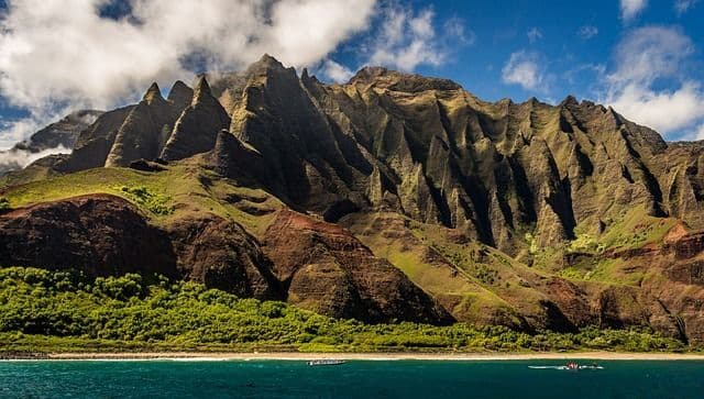Eine schroffe, grüne Bergkette mit scharfen Gipfeln erhebt sich steil aus dem blauen Ozean, dazu ein schmaler Strand und kleine Boote auf dem Wasser.