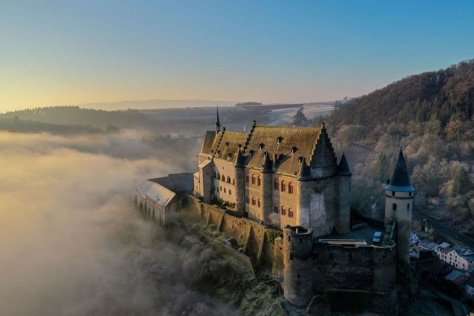 Una vista aerea di un grande castello di pietra su una collina, che si innalza sopra una fitta coltre di nebbia all'alba.
