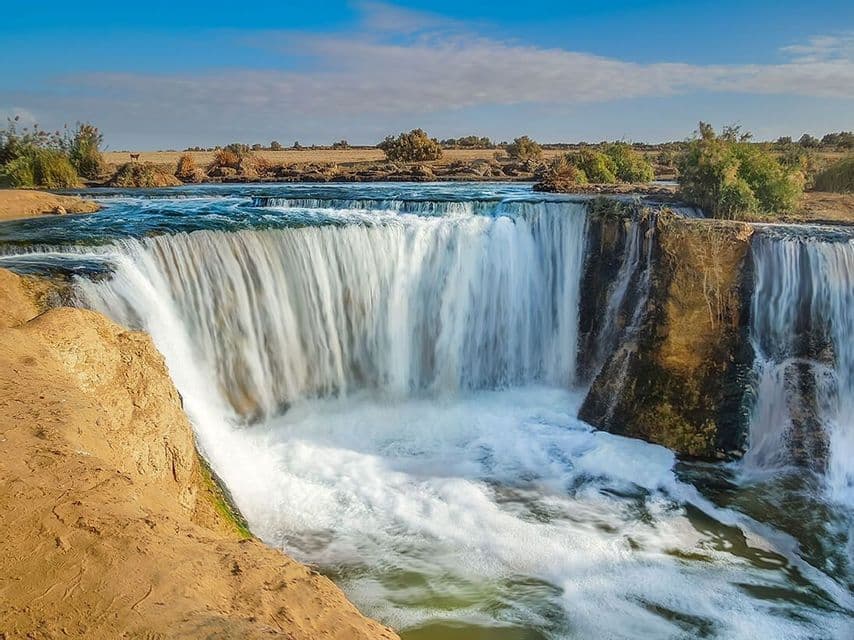 Un'ampia cascata scende da una scogliera rocciosa in un fiume impetuoso, con un paesaggio arido sullo sfondo sotto un cielo azzurro.