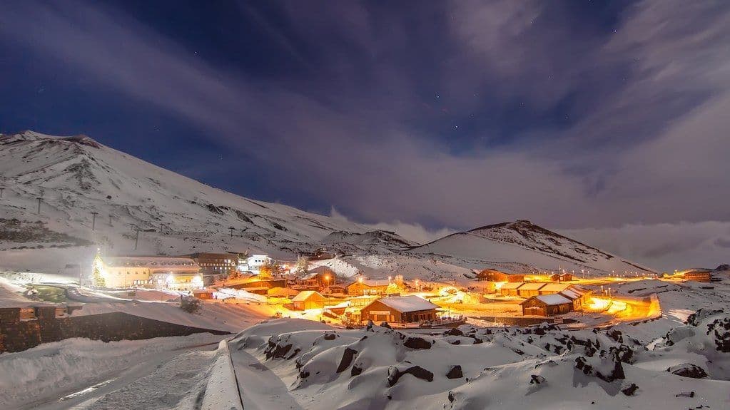 Un villaggio illuminato risplende in un paesaggio montano innevato di notte sotto un cielo nuvoloso.