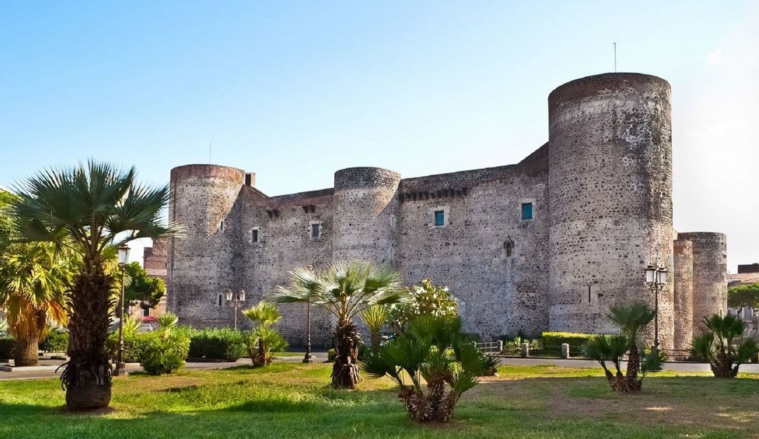 Un castello medievale in pietra con grandi torri rotonde, visto da un parco erboso con palme sotto un cielo sereno.