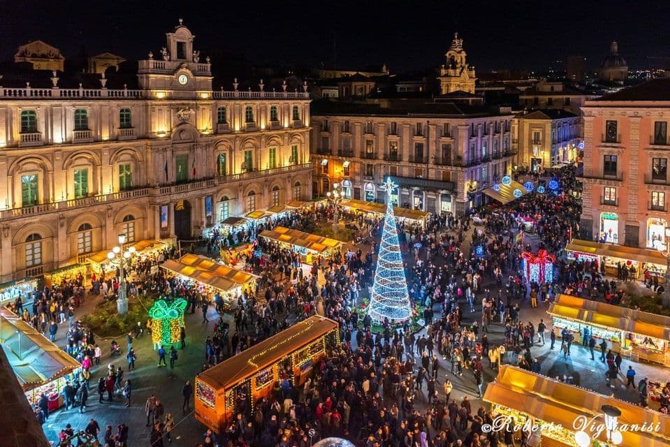 Veduta aerea di un affollato mercatino di Natale di notte, con un albero illuminato e bancarelle in una piazza storica.