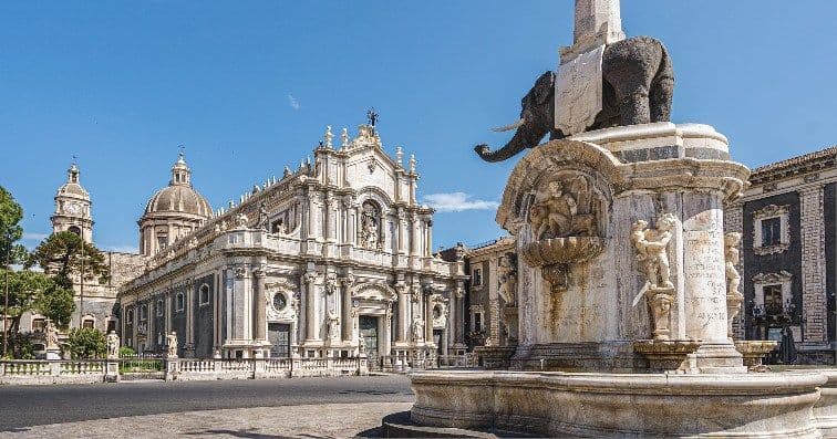 Una fontana in pietra con una statua di elefante si erge in un'ampia piazza di fronte a un'ornata cattedrale barocca sotto un cielo limpido.