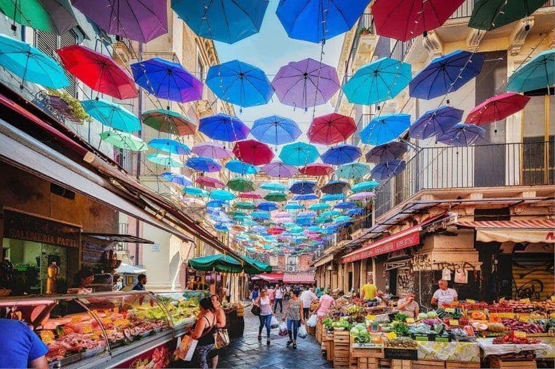Ombrelli colorati pendono su un vivace mercato di strada pieno di bancarelle di cibo fresco e persone che passeggiano.