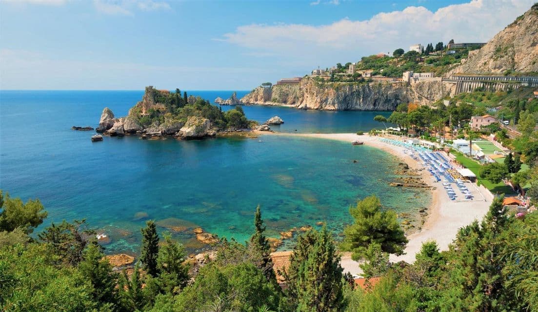 Vista dall'alto di una baia costiera con acqua cristallina color turchese, un isolotto roccioso e una spiaggia sabbiosa costellata di ombrelloni.
