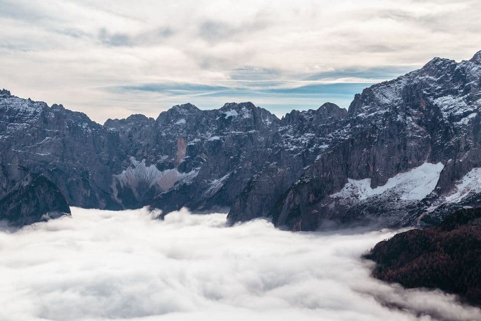Una catena montuosa aspra e innevata si erge sopra un denso mare di nuvole sotto un cielo parzialmente nuvoloso.