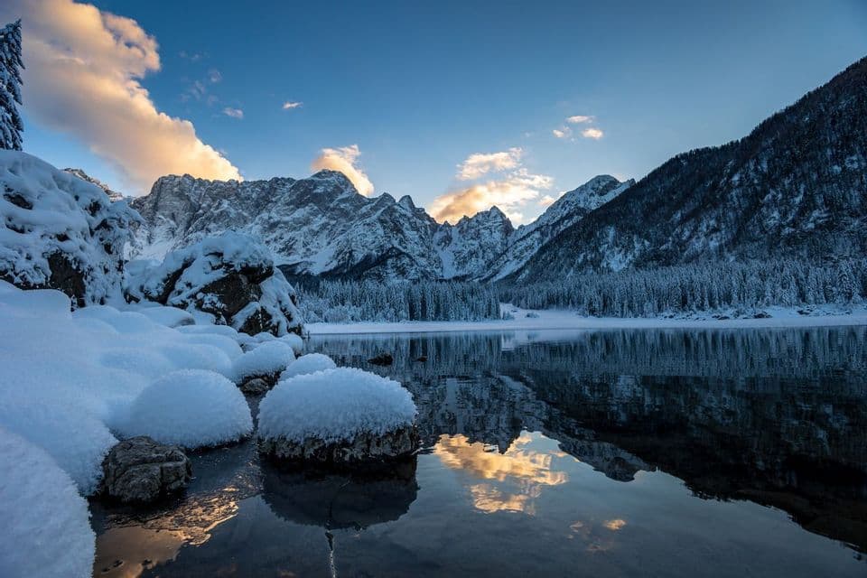 Montagne innevate e una pineta si riflettono in un lago calmo, con rocce coperte di neve in primo piano sotto un cielo blu.