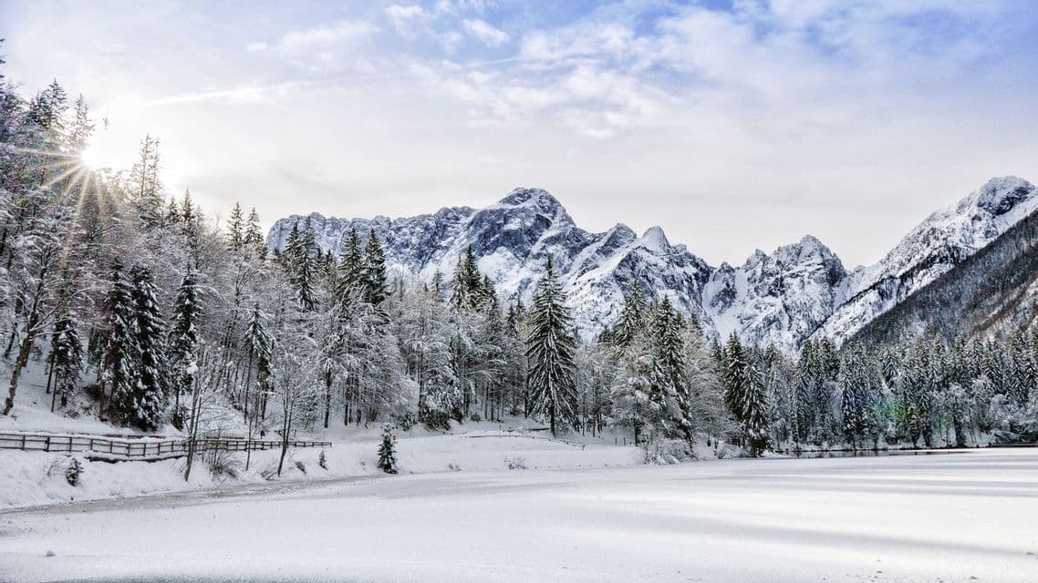 La luce del sole splende su una pineta coperta di neve e un lago ghiacciato, con una catena montuosa scoscesa sullo sfondo.