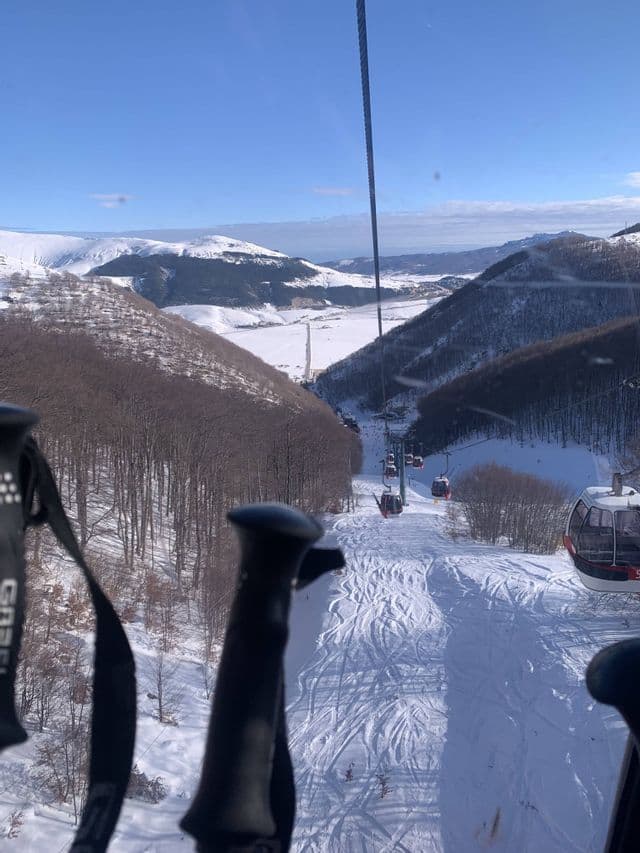 Una vista da una seggiovia che domina una valle innevata con piste da sci, alberi spogli e montagne sotto un cielo blu.