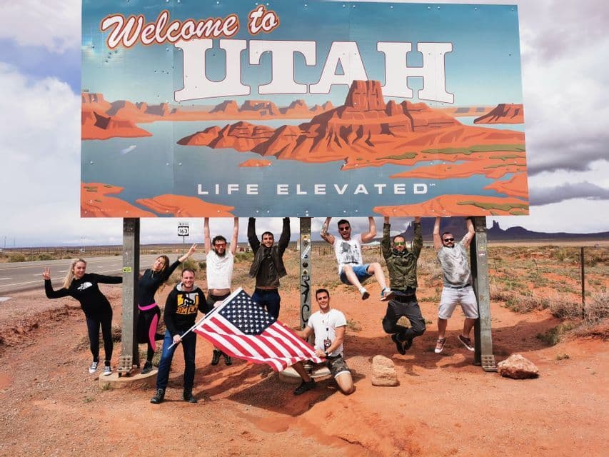 Members of a WeRoad group trip hang from and pose under a large 'Welcome to Utah' sign, with one person holding an American flag.